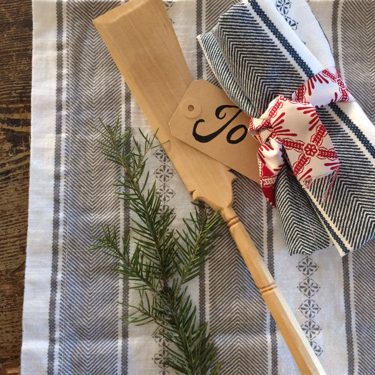 weiß-grau gestreiftes Geschirrtuch auf einem Holztisch, darauf hölzerner Pfannenwender, eine Stoffserviette mit dunkelgrauem Streifenmuster, die mit einem rot-weiß gemusterten Band zusammengebunden ist, Zweig von Nadelgrün liegt ebenfalls als Dekoration auf dem Tuch