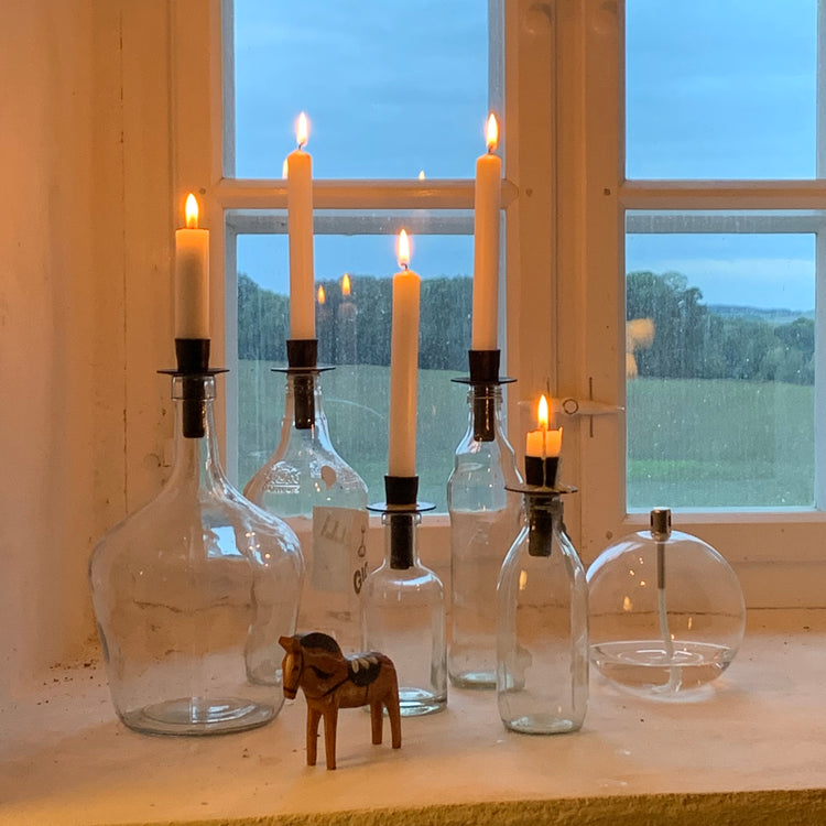 viele Glasflaschen mit schwarzen Kerzenhaltern Flaska in schwarz mit brennenden Kerzen in einem Fenster mit Ausblick in die Natur draußen