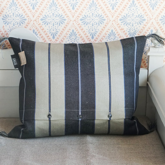 Rückseite des Leinenkissens breitgestreift - es sind fünf Streifen, außen je ein schwarzer dazwischen je einer in natur. Jeder Streifen hat mittig noch einmal einen schmalen Streifen, die schwarzen dicken Streifen haben einen Streifen in natur mit links und rechts einem gleichbreiten in blau, die breiten Streifen in natur haben einen schmalen blauen Streifen mit links und rechts je einem gleichschmalen in schwarz. An den Ecken sind Fransenbommeln aus Leinenfasern. Im unteren Drittel Knopfleiste.