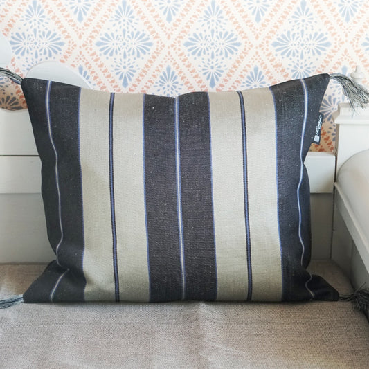 Vorderseite des Leinenkissens breitgestreift - es sind fünf Streifen, außen je ein schwarzer dazwischen je einer in natur. Jeder Streifen hat mittig noch einmal einen schmalen Streifen, die schwarzen dicken Streifen haben einen Streifen in natur mit links und rechts einem gleichbreiten in blau, die breiten Streifen in natur haben einen schmalen blauen Streifen mit links und rechts je einem gleichschmalen in schwarz. An den Ecken sind Fransenbommeln aus Leinenfasern