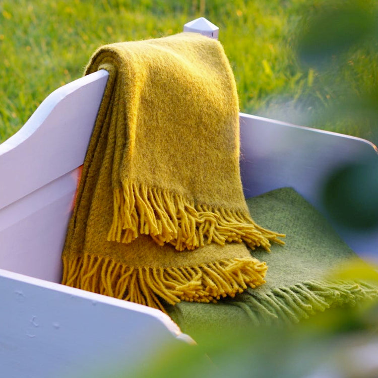 gelbe Gotlandwolldecke hängt über dem Fußende eines grauen Holzbettes, das in einer Wiese steht
