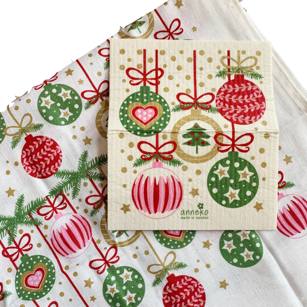 Schwammtuch auf einem Teil des Geschirrhandtuchs liegend, fünf Weihnachtskugeln mit rot/grün/golden Mustern mit roten Aufhängungen sind abgebildet auf dem Schwammtuch. Das gleiche Motiv auf dem Geschirrtuch