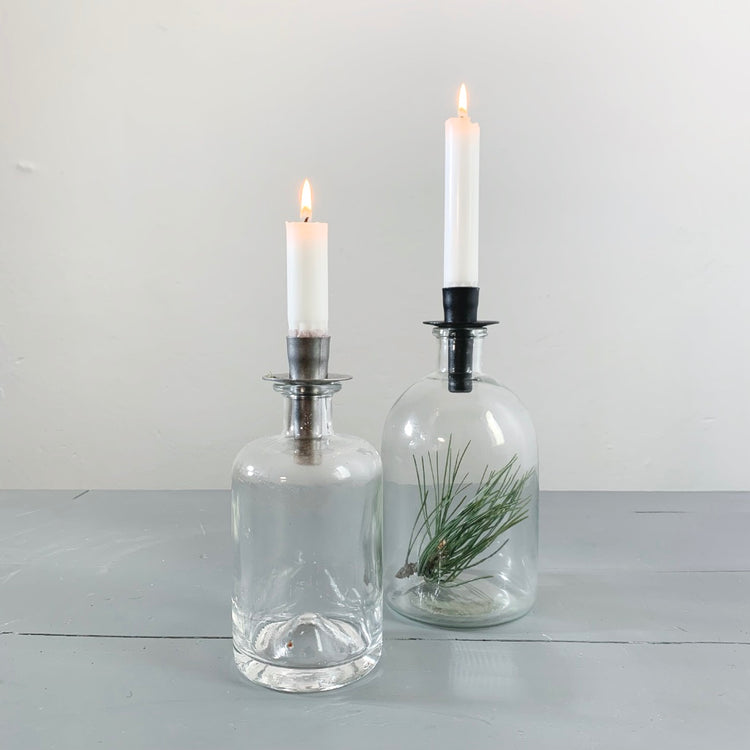 zwei Glasflaschen mit je einem Kerzenhalter Flaska in schwarz bzw. unbehandelt. Eine Flasche enthält einen grünen Kiefernzweig