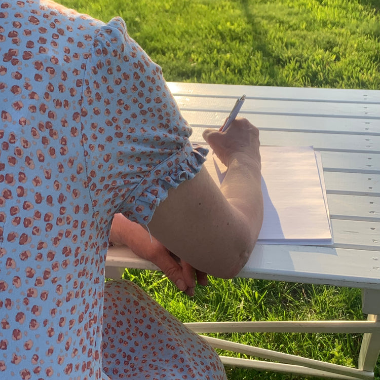 Frau von hinten, sitzend an Gartentisch in Abendsonne und einen Brief schreibend