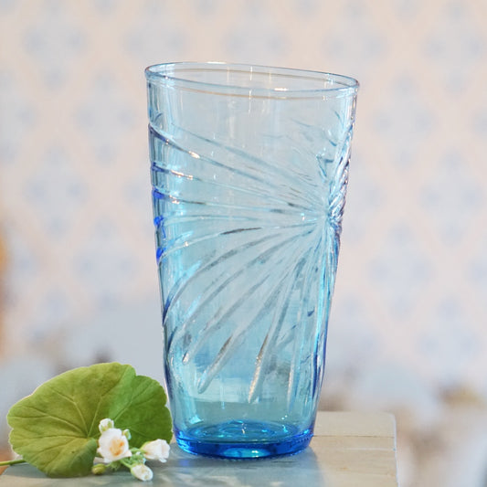 hellblaue Vase aus Glas mit eingearbeitetem Strahlenmuster, am Fuß liegt eine Pelargonienblüte