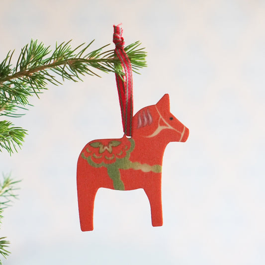 rotes, ausgesägtes Dalapferd mit Blumenmuster mit einem schmalen Webband in rot und grün an Tannenzweig hängend