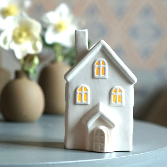 Kleines Haus aus weißer Keramik, das von innen leuchtet, im Hintergrund weiße Christrosen