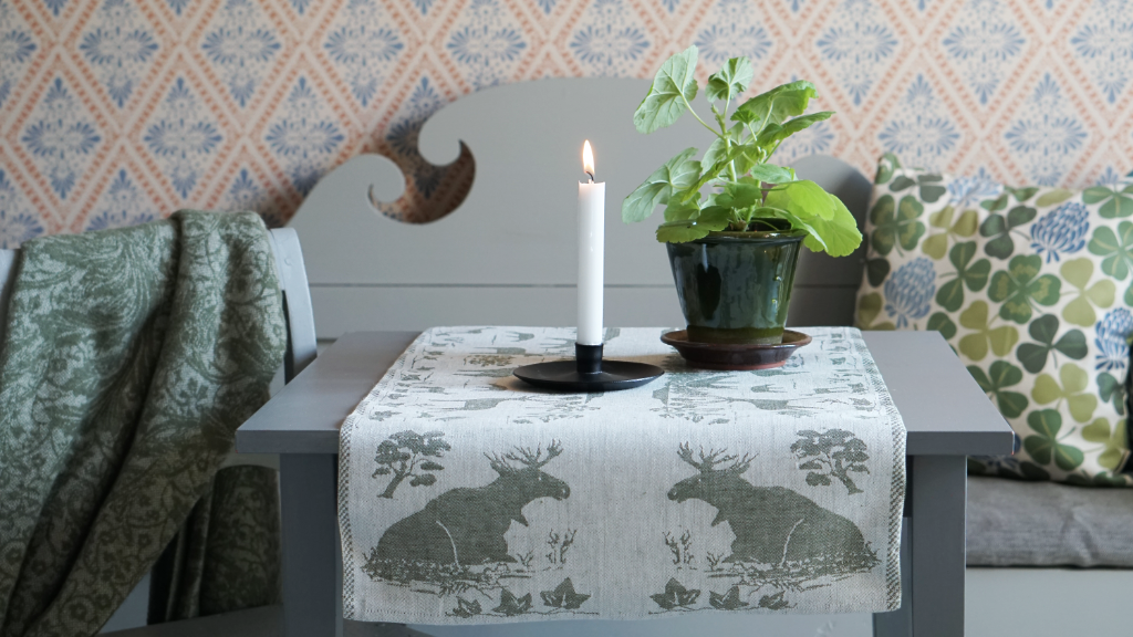 Tisch mit Läufer in Leinen vor Küchensofa, auf dem Tisch Kerze und Blumentopf mit Pelargonie alles in grün gehalten