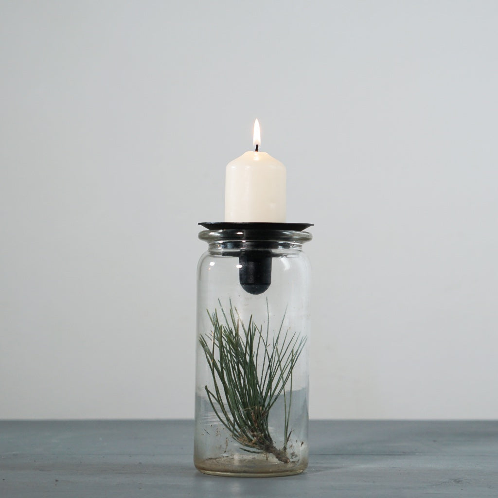 Glas mit Kiefernzweig obenauf der Kerzenhalter Burk in schwarz mit brennender Blockkerze
