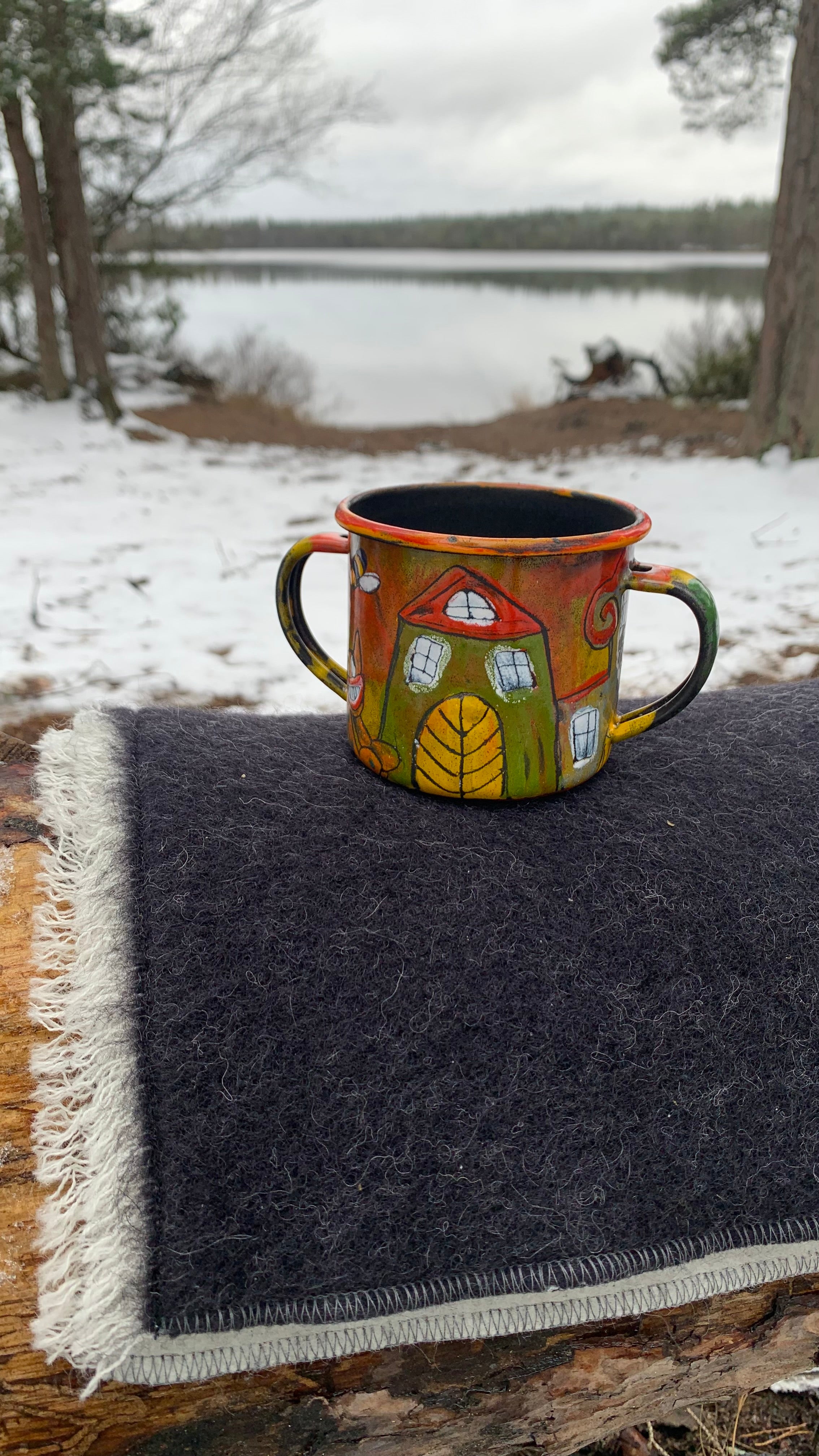Sitzunterlage aus Wolle anthrazit/weiß auf Baumstamm am See mit bunter Emailletasse im Winter
