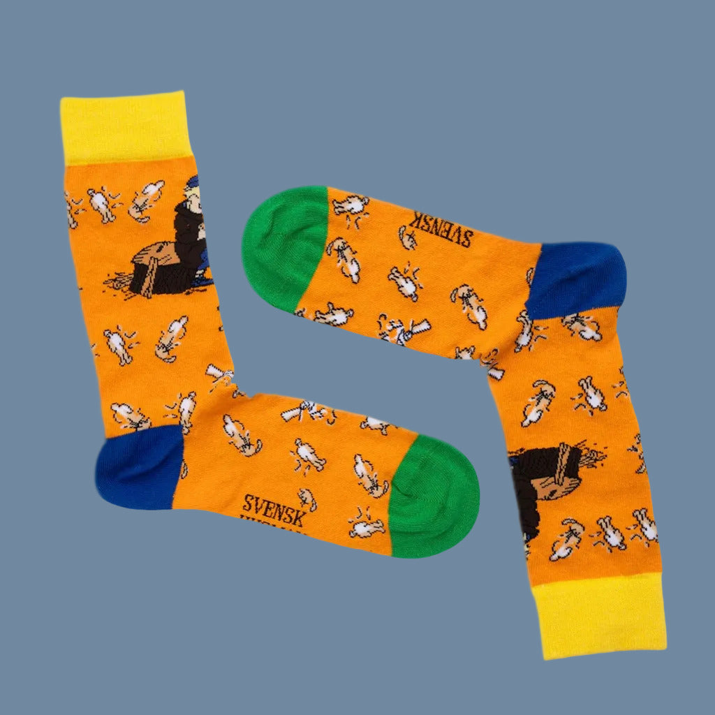 Orange Socken mit Motiv Michel macht Holzmännchen mit grüner Spitze, gelbem Bündchen und blauer Ferse vor mysblauem Hintergrund