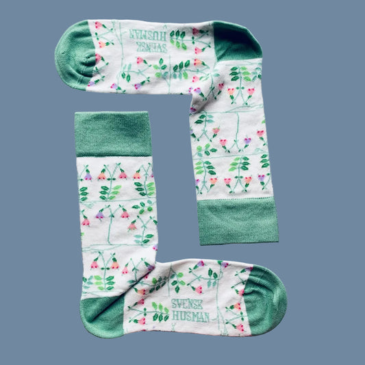 Weiße Socken mit Linnea Blüten und Blättern und grüner Ferse und Spitze vor mysbluem Hintergrund