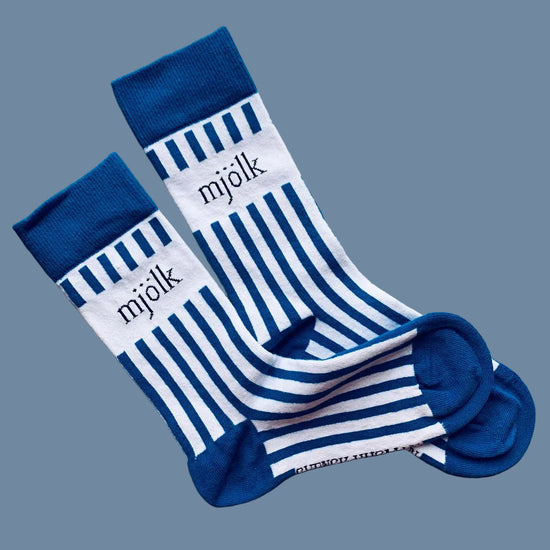 Weiße Socken mit blauen Streifen und Aufschrift Mjölk mit blauer Ferse und Spitze vor mysblauem Hintergrund
