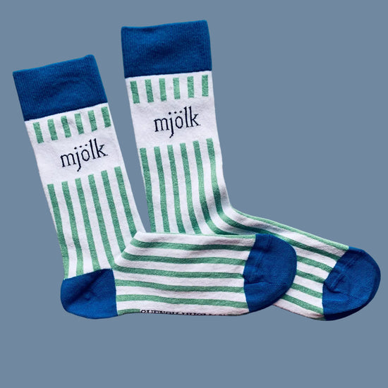 Weiße Socken mit grünen Streifen und Aufschrift Mjölk mit blauer Ferse und Spitze vor mysblauem Hintergrund
