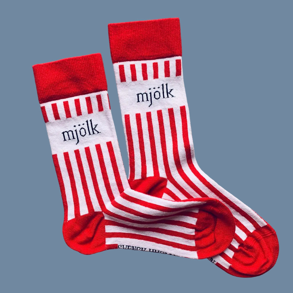 Weiße Socken mit roten Streifen und Aufschrift Mjölk mit roter Ferse und Spitze vor mysblauem Hintergrund
