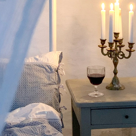 Interiorbild mit Ecke eines Bettes in dem Set aus Kopfkissen und Bezug mit Muster Destiny blau liegen, daneben ein Nachttisch mit Kerzenleuchter und Weinglas